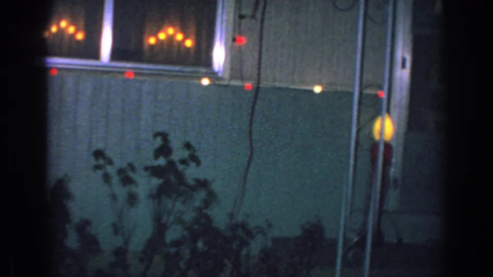 1962 Gary, Indiana home-movie frame showing part of the decorated house exterior with glowing window lights, colored bulbs, shrubs, and a tall red holiday decoration near the wall.