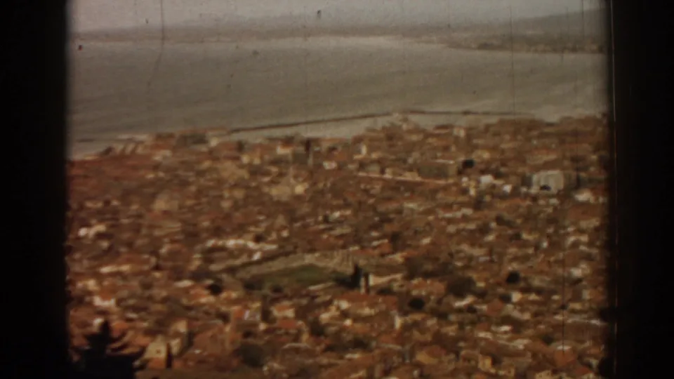 Archival 8mm frame showing Izmir's dense rooftops below the bay, with warm color, grain, and a dark window edge.