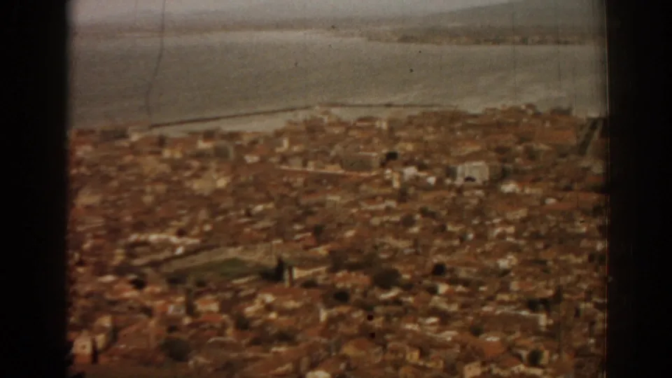 Window-framed 1962 home-movie view over Izmir rooftops and waterfront, with visible 8mm grain and muted warm tones.