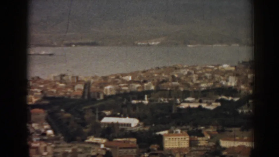 1962 Izmir home-movie frame with foreground buildings, dark parkland, bay traffic, and mountain slopes beyond.