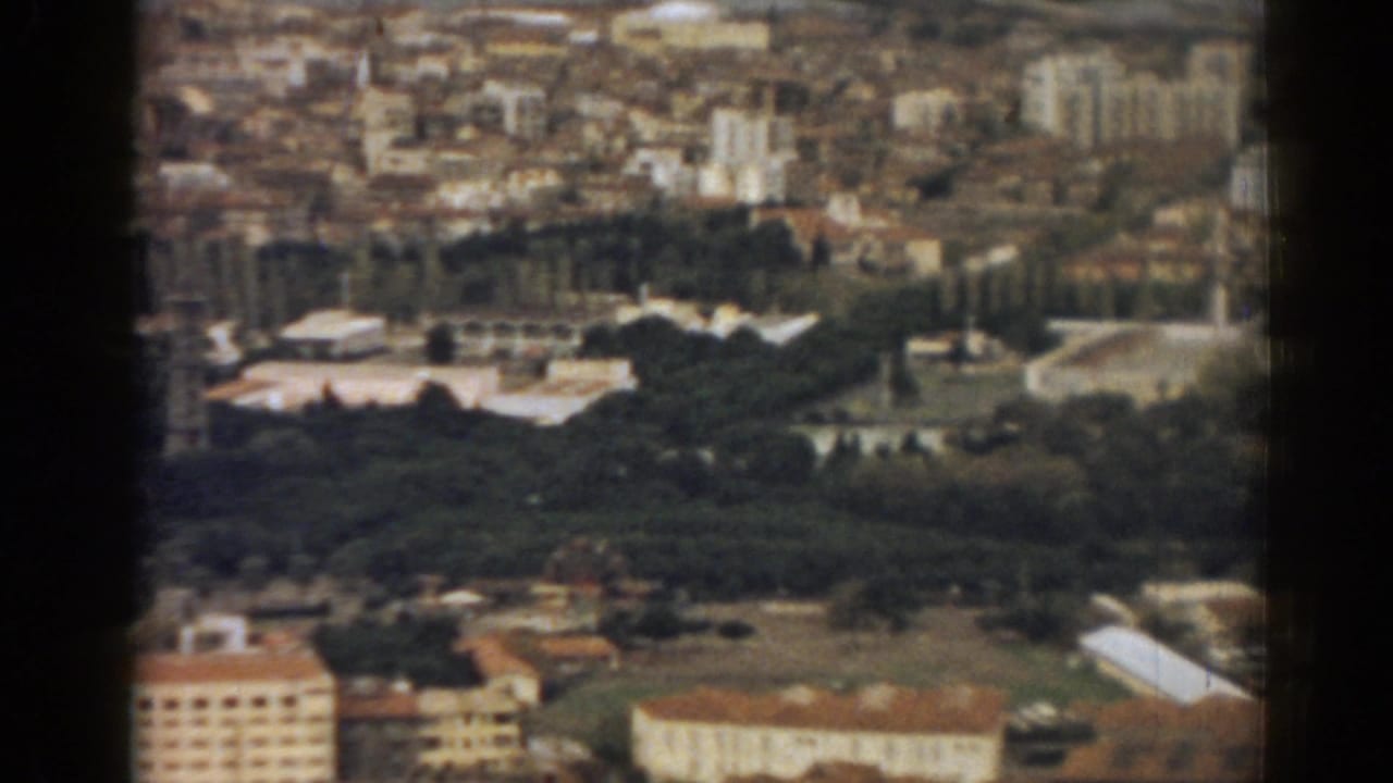 1962 Izmir aerial frame showing dense buildings, green tree cover and a mosque minaret amid the cityscape.