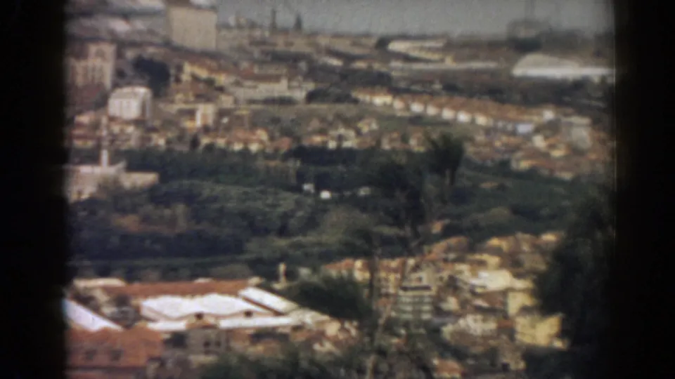 Soft 8mm frame of 1962 Izmir from an overlook, with foreground rooftops, dark tree cover, dense buildings, and coastal water beyond.