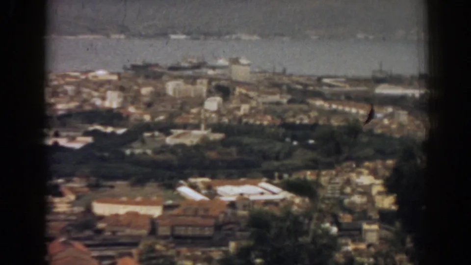 Wider 1962 Izmir overlook showing a built-up valley, terracotta roofs, tree cover, bay water, and distant ships under hazy light.