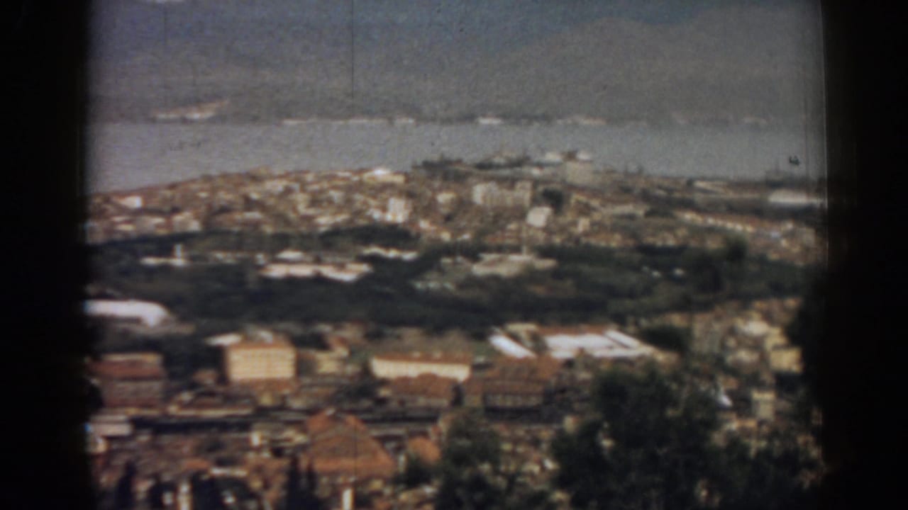 High-angle 1962 home-movie frame of Izmir with dense waterfront buildings, shoreline water, and muted mountains in the distance.