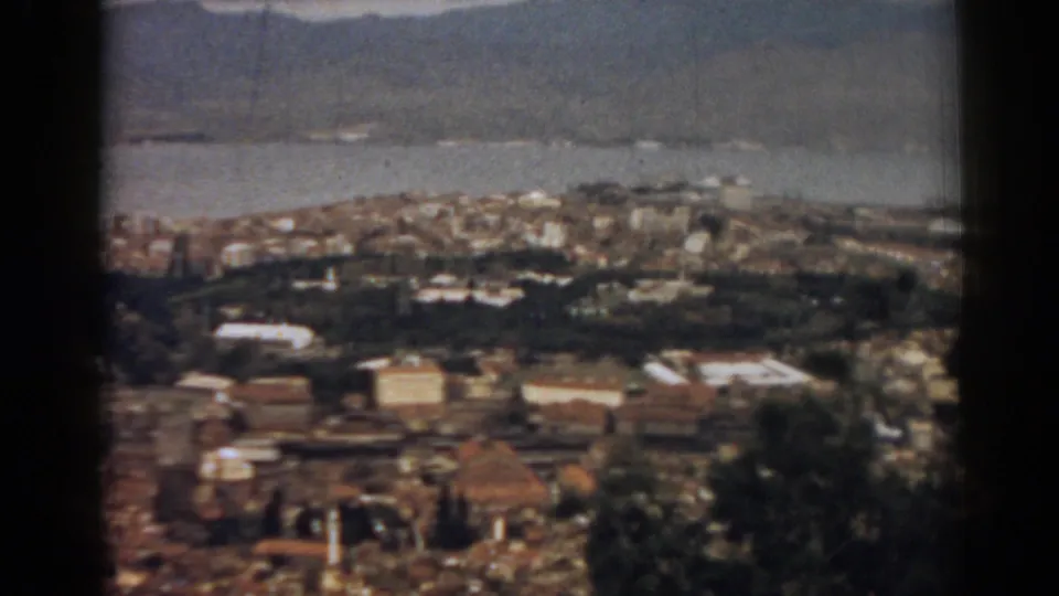 Soft-focus 8mm frame of Izmir from above, with coastal water behind low-rise buildings, greenery, and hazy mountain forms.
