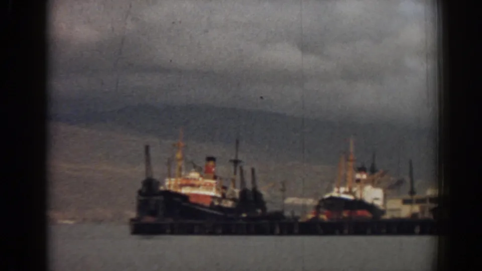 1962 Izmir harbor view with docked ships at a pier, calm water, muted sky, and a hazy mountain backdrop.