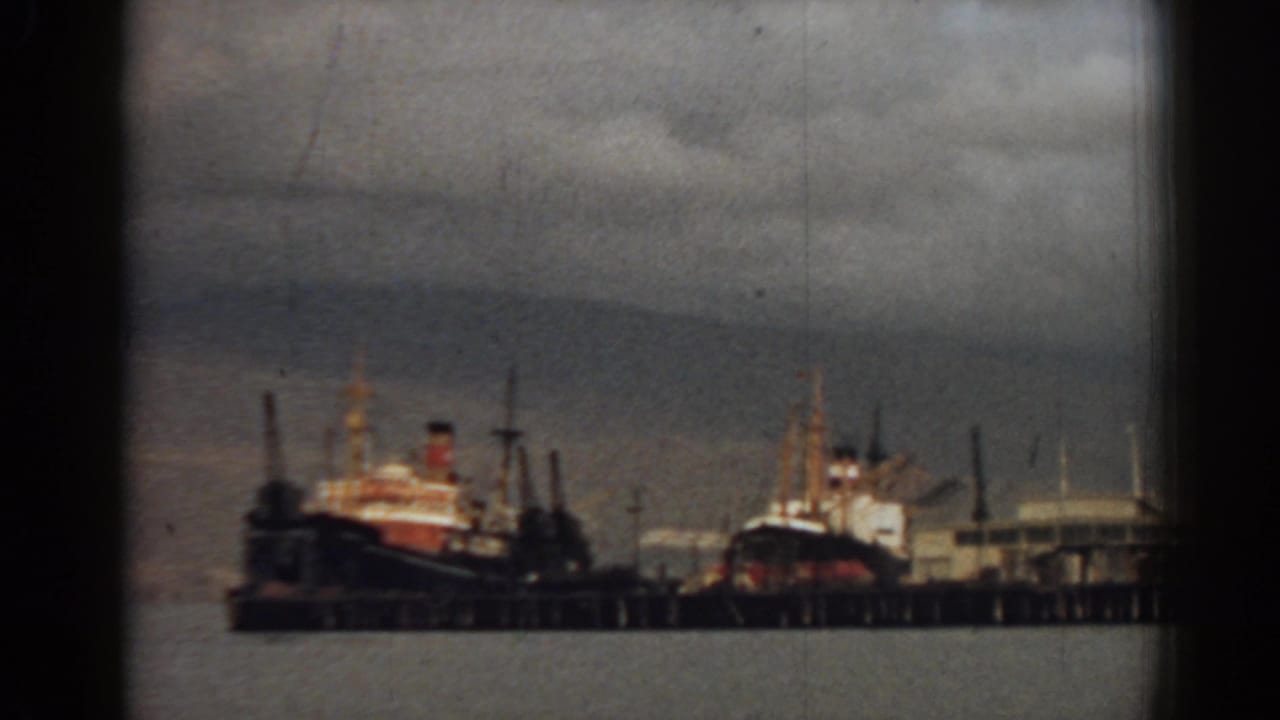 Docked ships along a pier under a cloudy sky, seen across harbor water.