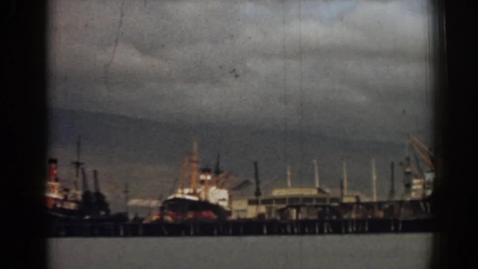 1962 Izmir port view with a dark-hulled ship, pier, cranes, harbor water, and muted cloudy sky on faded 8mm film.