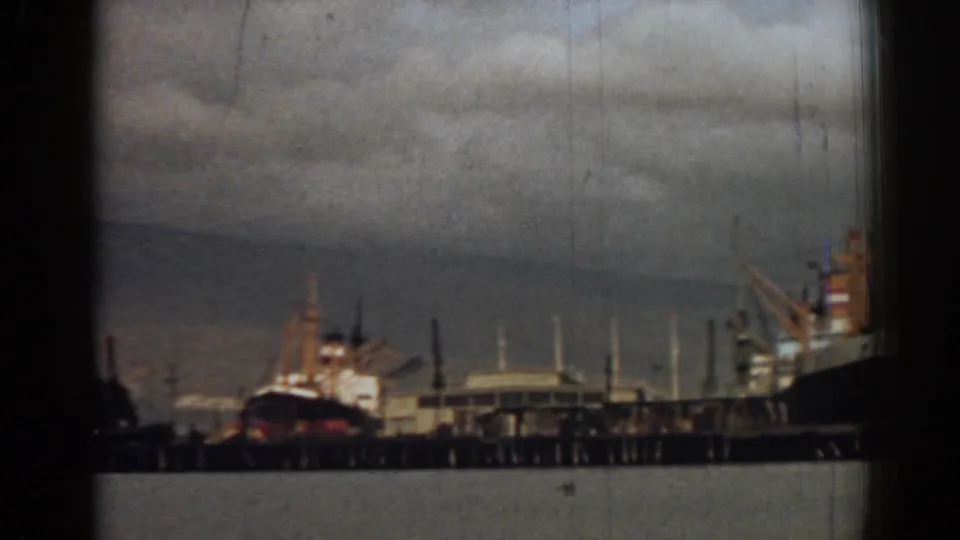 Final 1962 8mm frame of the Izmir waterfront, showing docked ships, a pier, cranes, water, and cloudy sky.