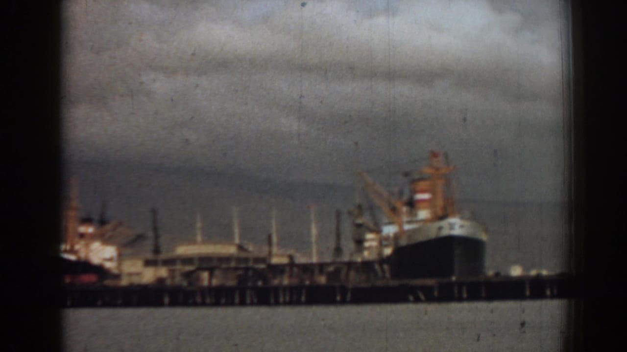 Large dark-hulled ship docked beside port cranes and buildings.
