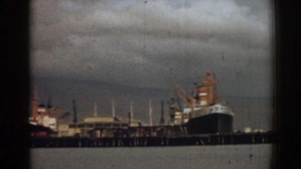 Color 8mm frame from Izmir, Turkey, showing a dark cargo ship at the pier with cranes and harbor buildings in the background.