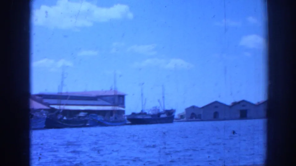 Blue-shifted 8mm frame of the Izmir harbor area, with small boats, a waterfront building, and warehouse-like structures across the water.