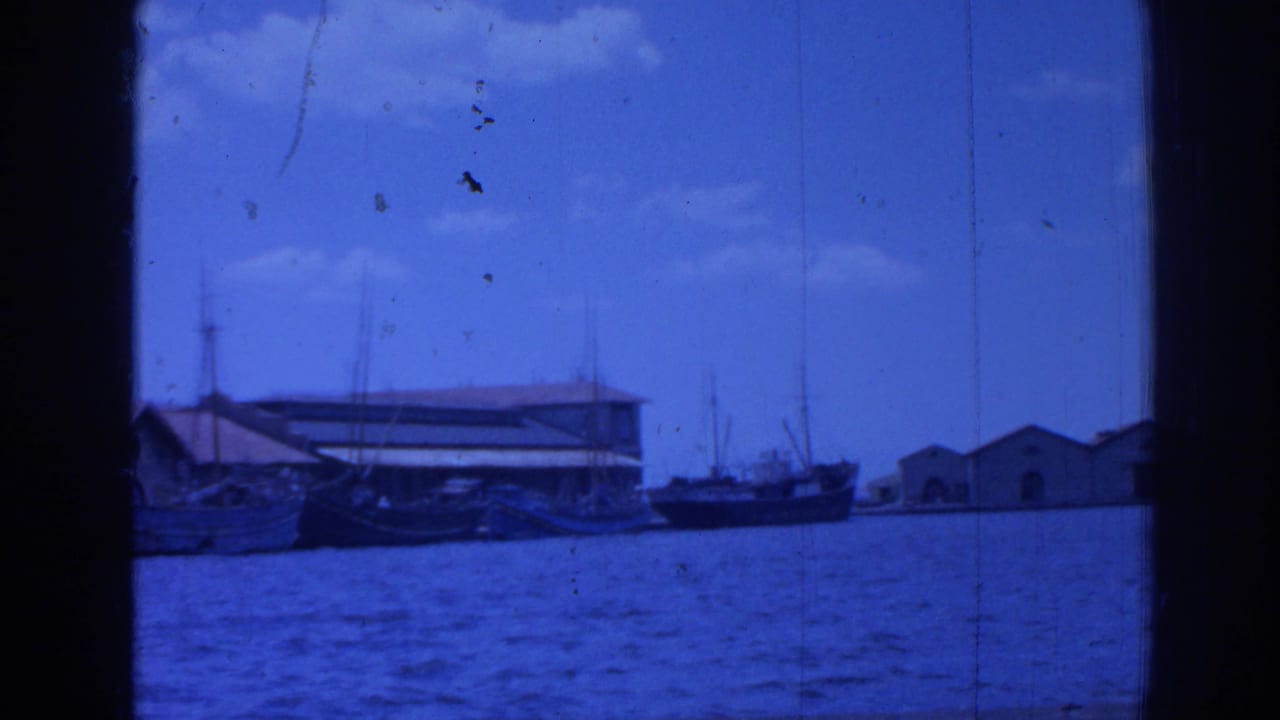 1962 Izmir harbor view across blue water, with small boats, pale waterfront buildings, masts, and dark 8mm frame borders.