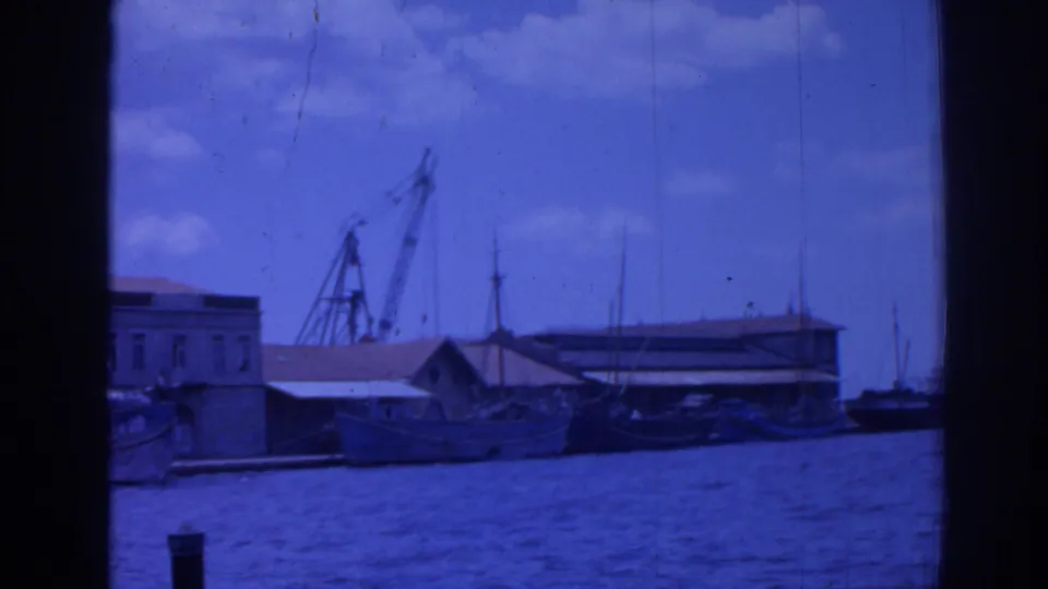Blue-tinted 1962 Izmir harbor frame showing a crane, moored boats, low waterfront buildings, masts, and bright clouds.
