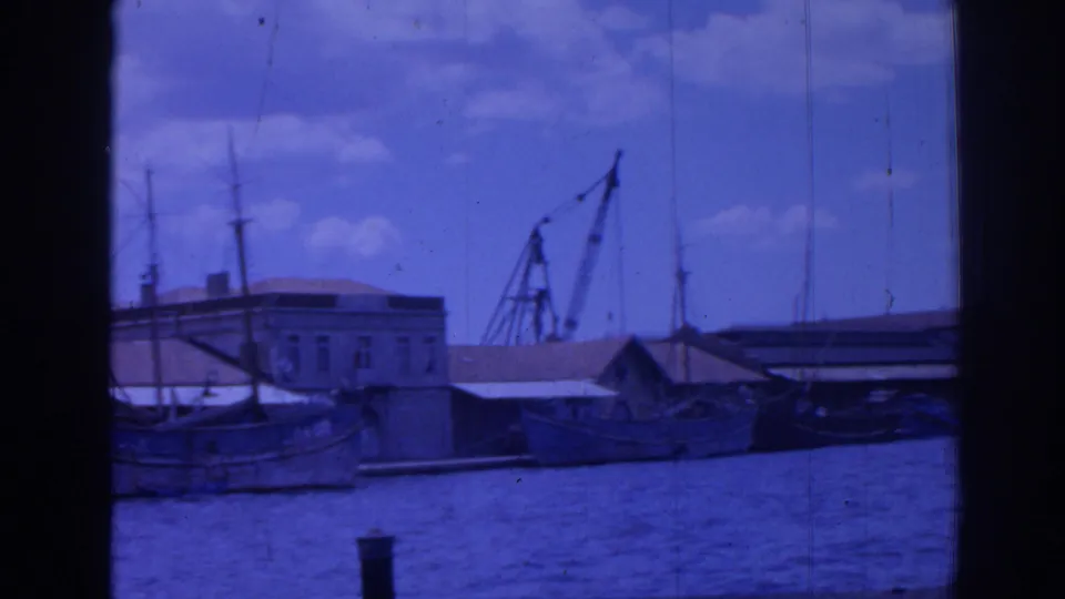 1962 Izmir harbor view with boats along the quay, a crane behind low buildings, masts, blue water, and 8mm gate borders.