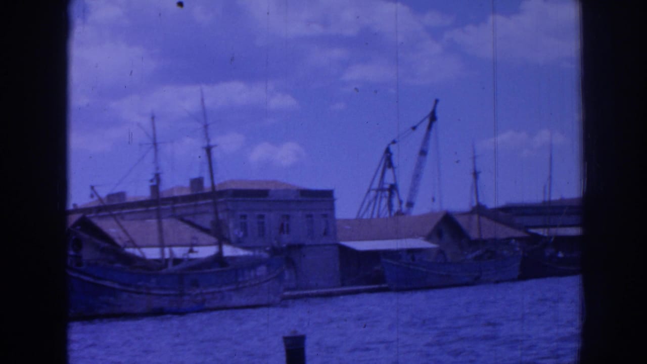 1962 Izmir harbor view with boats along the quay, waterfront buildings, mast lines, and a dockside crane under a cloudy sky.