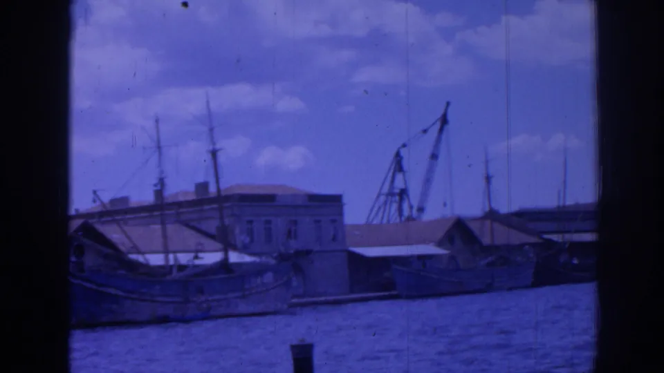 1962 Izmir harbor view with boats along the quay, waterfront buildings, mast lines, and a dockside crane under a cloudy sky.