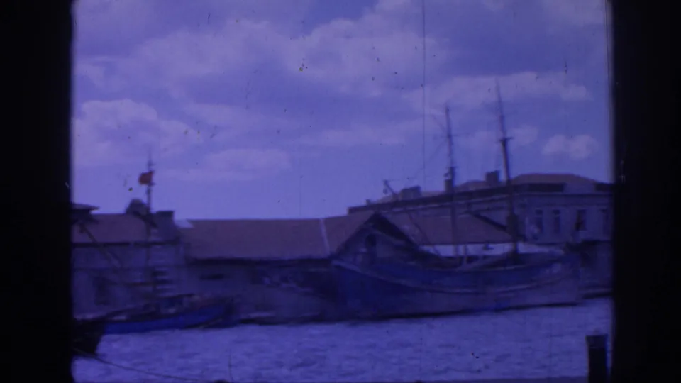 Izmir dockside scene from 1962 with a boat hull in the foreground, low waterfront structures, masts, and blue-cast home-movie grain.