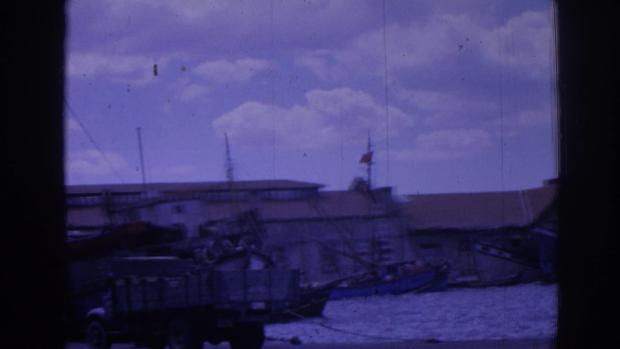 Harbor buildings, moored boats, water, and a truck seen through a dark 8mm frame edge.