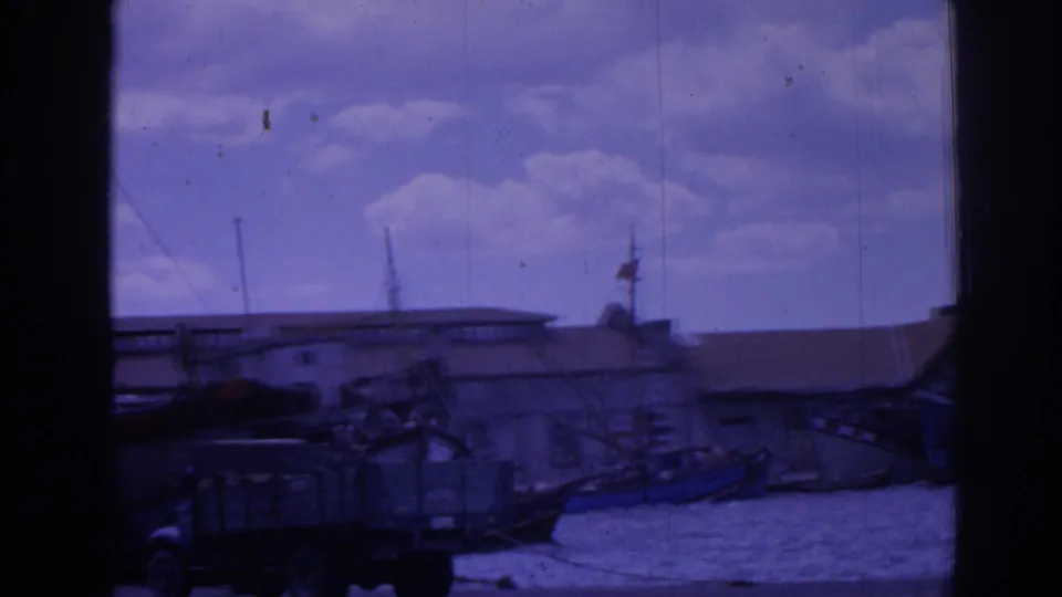 1962 Izmir harbor view with dockside truck, low buildings, water, masts, and boats under a cloudy sky.