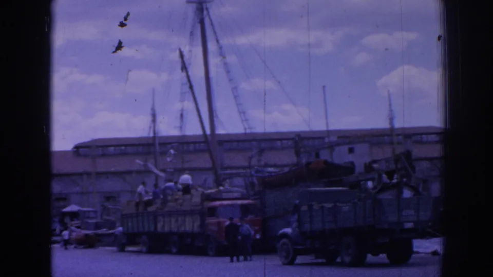 1962 Izmir waterfront showing workers near trucks and cargo, with moored boats and tall masts against harbor buildings.