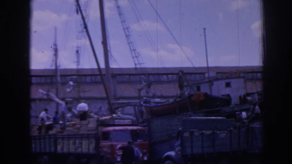 1962 Izmir harbor frame with workers near loaded trucks, a small boat, tall masts, water, and low waterfront buildings.