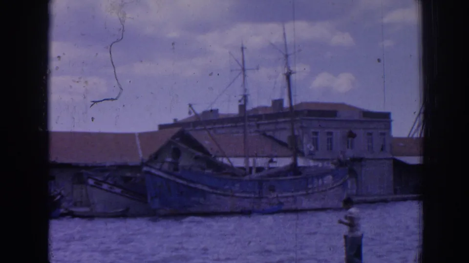 1962 Izmir waterfront with a weathered wooden boat, masts, tiled roofs, low buildings, water and cloudy sky.