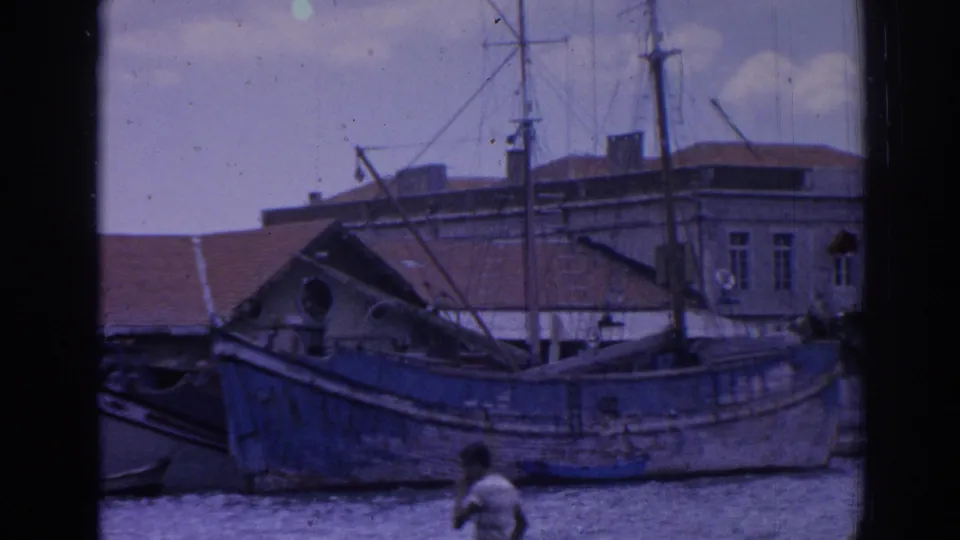 Izmir harbor frame with a blue wooden vessel, waterfront buildings, masts and a man in a light shirt walking near the pier.