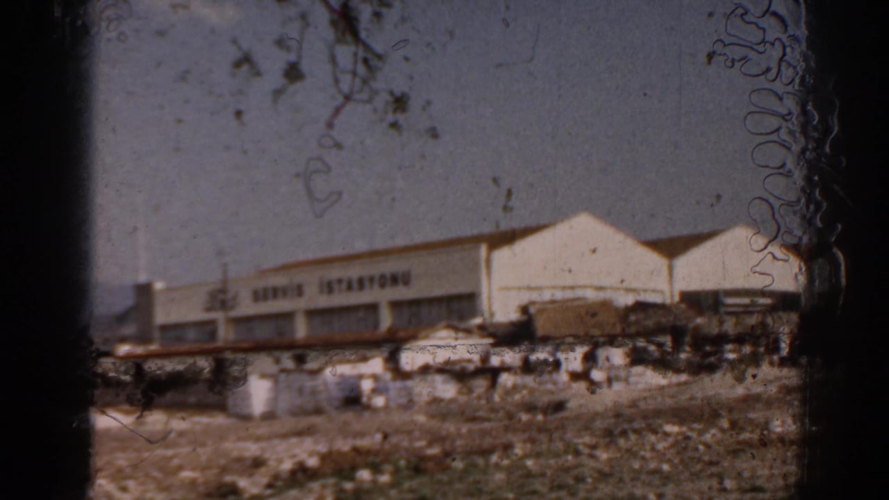 1962 Izmir home-movie frame showing a white Ford service station with Turkish signage beyond rough ground and small roadside structures.
