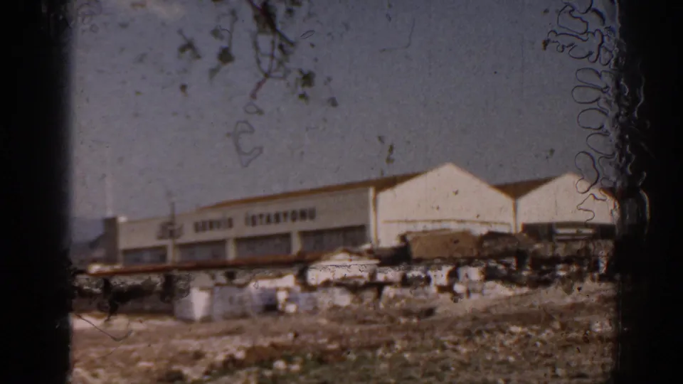 1962 Izmir home-movie frame showing a white Ford service station with Turkish signage beyond rough ground and small roadside structures.