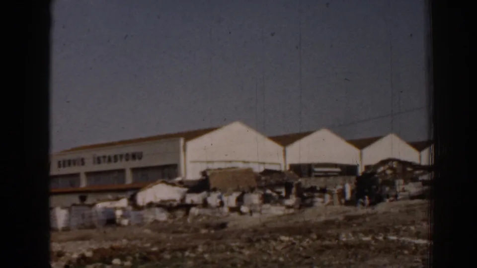 1962 Izmir home-movie frame showing the gabled white service station, partial Turkish signage, scattered containers, and roadside activity.