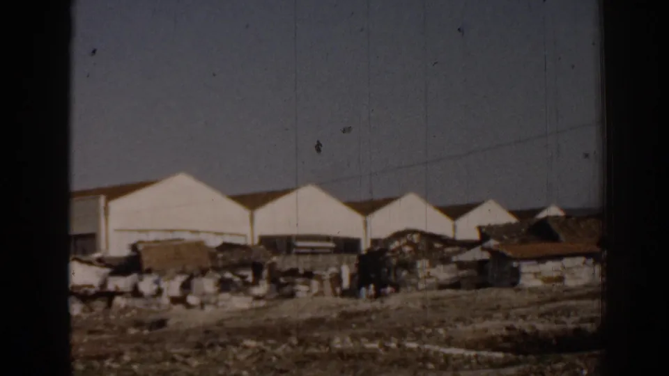 Late frame from the 1962 Izmir clip showing the white service station complex, open roadside ground, scattered materials, and distant figures.