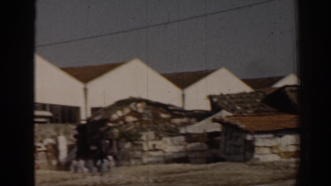 Low shacks and rubble in front of large white gabled buildings, with small figures nearby.