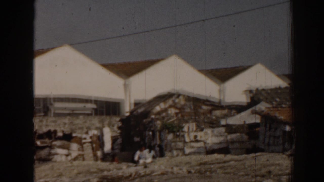 1962 Izmir frame with distant people near dilapidated dwellings, set against larger white modern-looking buildings.