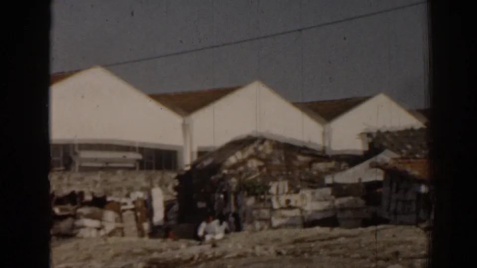 1962 Izmir frame with distant people near dilapidated dwellings, set against larger white modern-looking buildings.