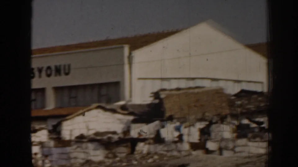 1962 Izmir home-movie frame showing a white building with partial visible lettering behind patched roofs and rough walls.