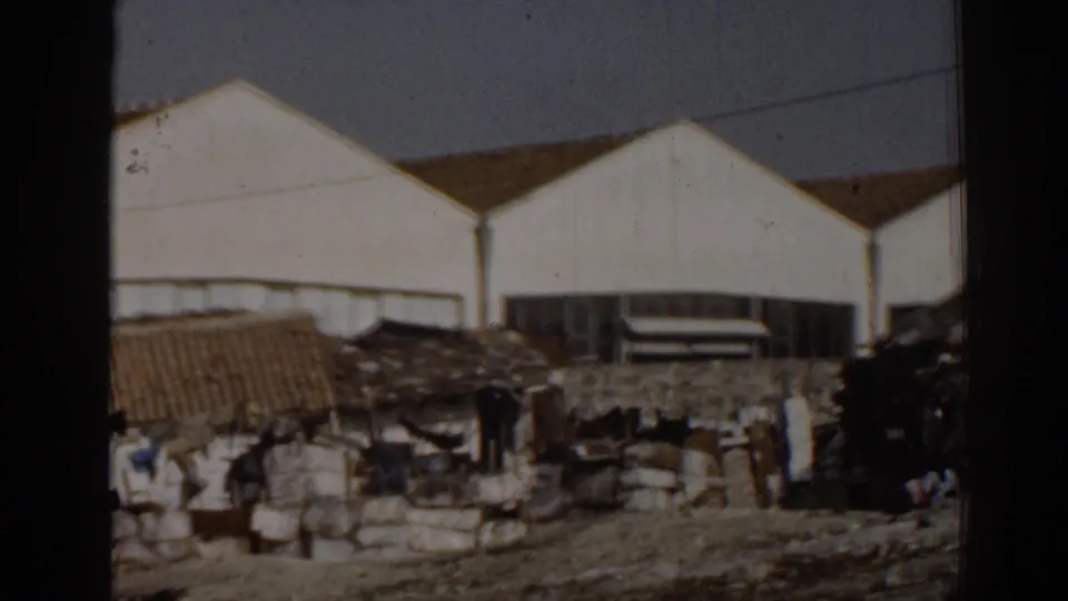 1962 Izmir 8mm view of factory-like buildings, low improvised structures, rubble, and a distant person at frame edge.