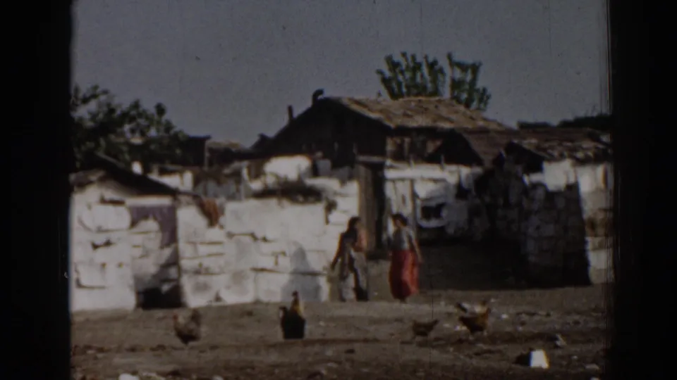 1962 Izmir frame showing women in a debris-strewn yard with chickens, low walls, shacks, and trees in the background.
