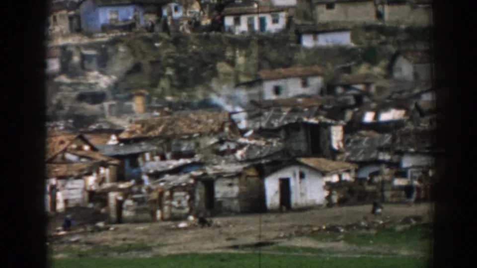 1962 Izmir home-movie frame showing a dense hillside settlement with modest dwellings, unpaved ground and small figures in the distance.