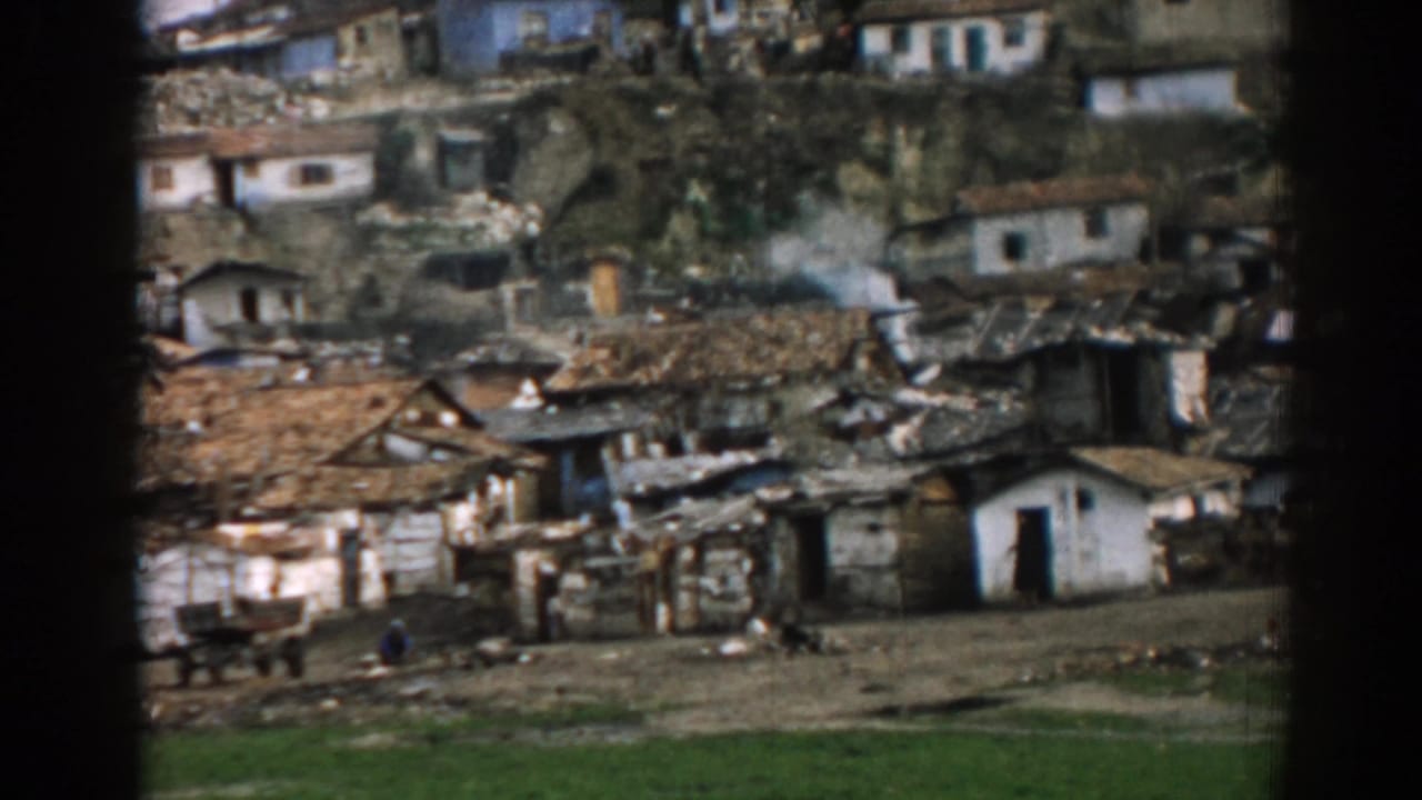 Hillside homes with patched roofs, a white doorway, rough yards and smoke rising nearby.