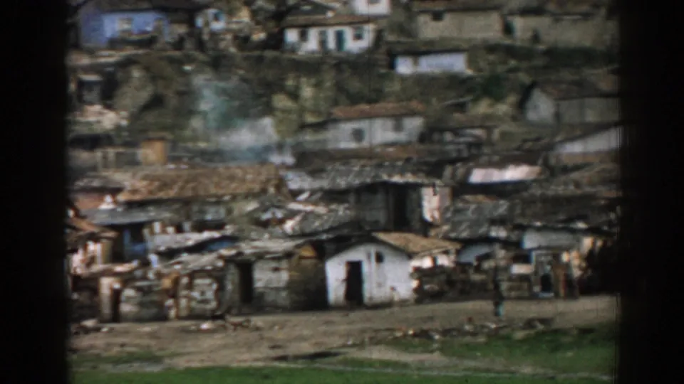 1962 Izmir frame showing a compact hillside village, smoke, rough yards, modest dwellings and distant residents outdoors.