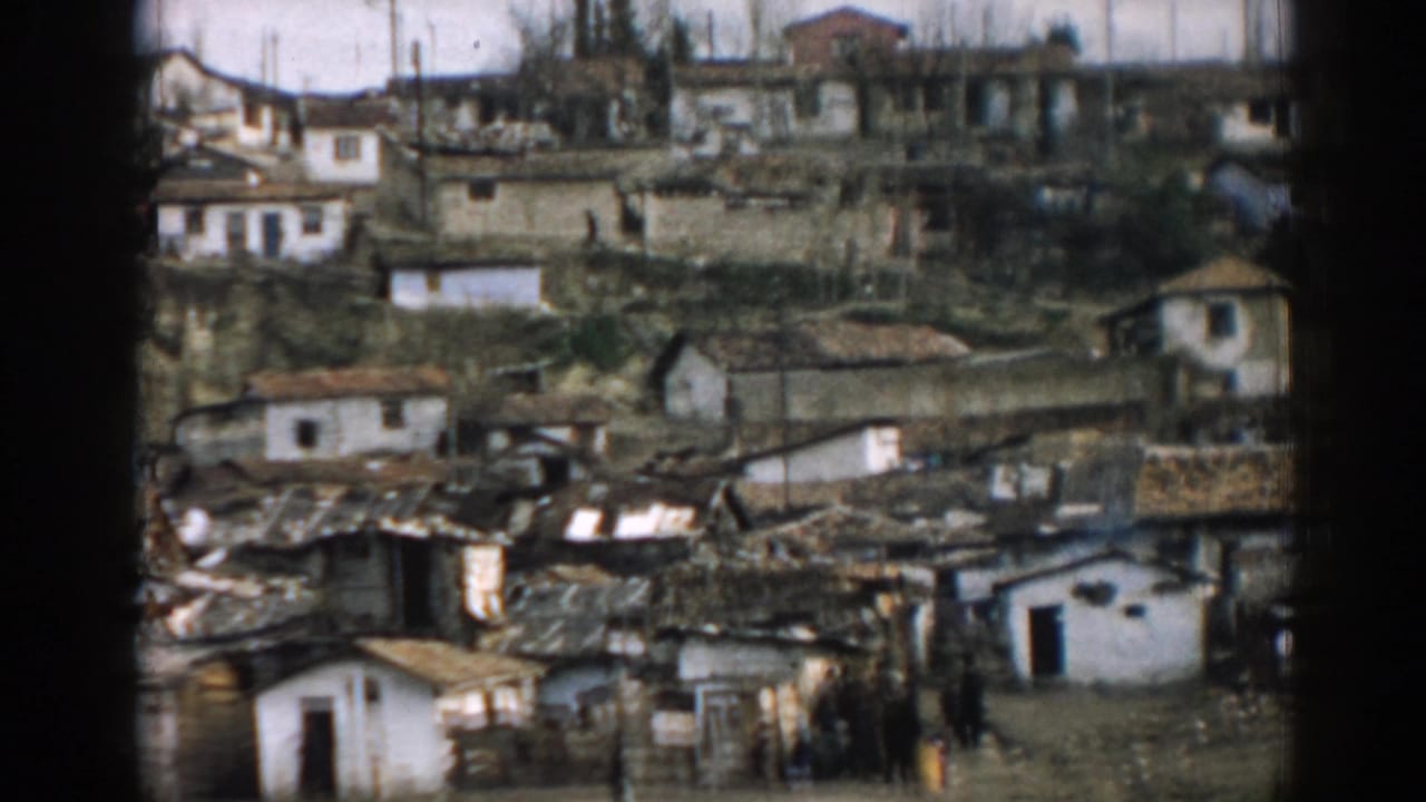Damaged hillside settlement with roof debris, white houses, and people gathered below.