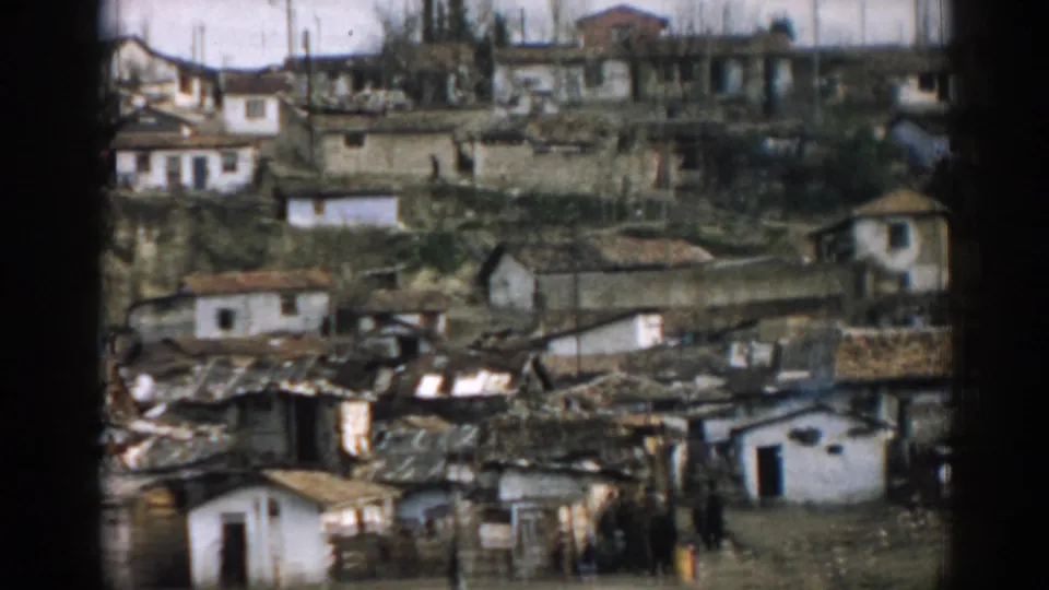 View across an Izmir hillside settlement showing burned rooflines, damaged homes, and residents in the foreground.