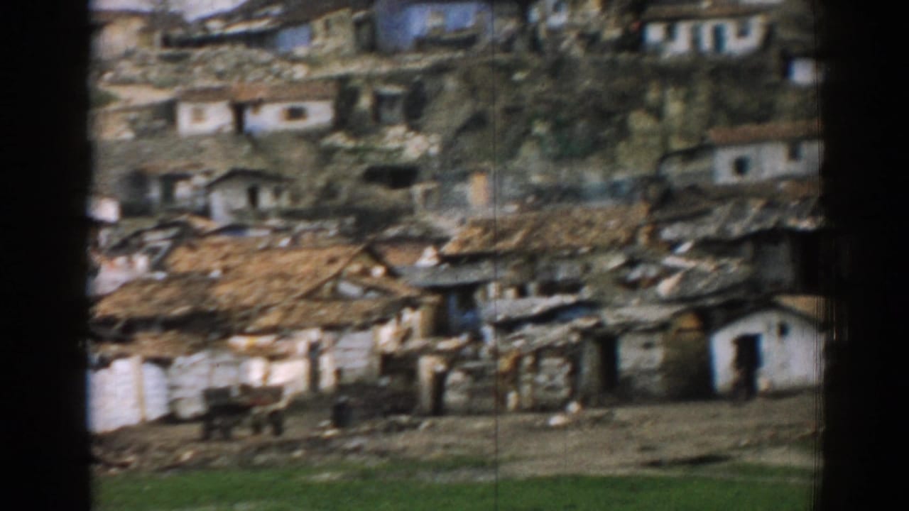 Hillside settlement with modest houses, rough ground, and a green grass strip in front.