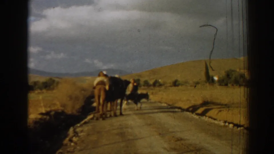 1962 Izmir, Turkey 8mm frame of donkey riders receding along a rural road, with golden hills, distant trees, and film scratches visible.