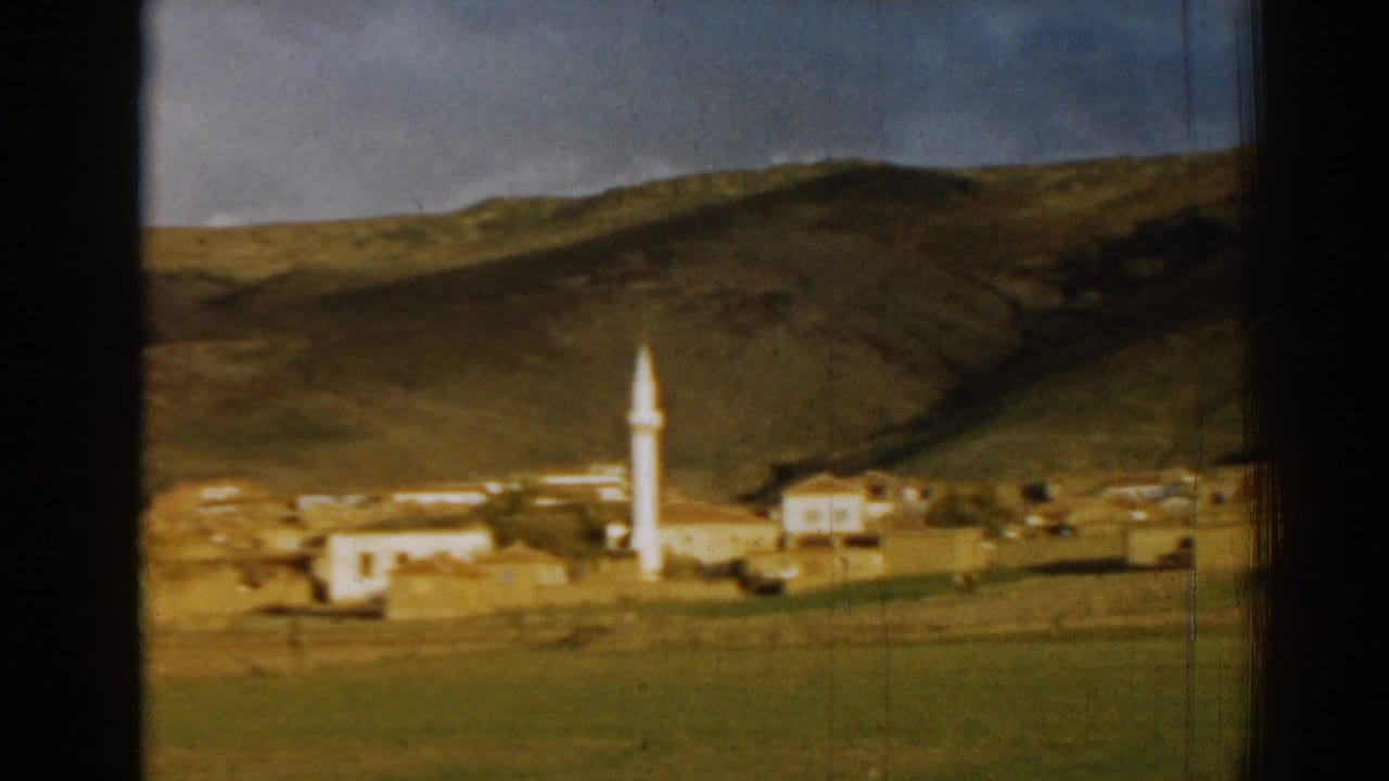 1962 8mm view near Izmir, Turkey, showing a small hillside settlement with a white minaret, low buildings, grass, and film-frame edges.