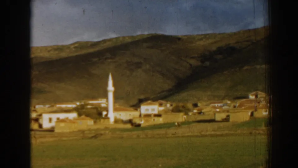 1962 Izmir-area home-movie frame of a rural settlement with a white minaret, pale buildings, grassy land, and hills in the background.