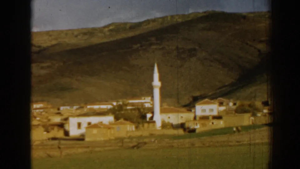 1962 8mm view near Izmir, Turkey, with the pan shifted across a hillside village, mosque minaret, low buildings, and open field.