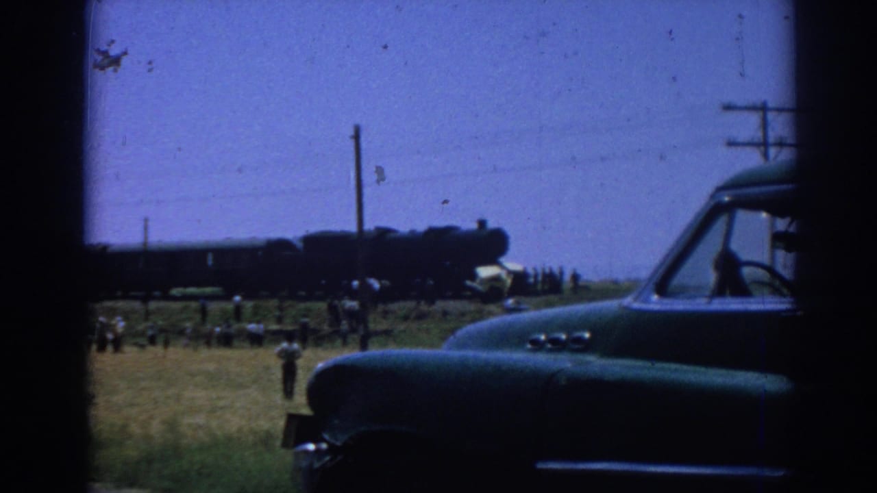 Green car in foreground with a dark steam train and scattered onlookers beyond a grassy field.
