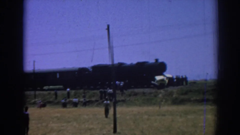 Wide 8mm view near Izmir showing a stopped steam train across an open field, with scattered onlookers and utility poles in frame.